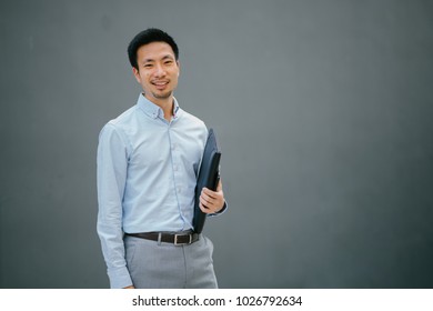 Portrait Of A Young, Chinese, Pan Asian Business Man Standing Against A Plain Grey Background And Smiling While He Holds His Leather Folio. He Is Well-dressed In Business Casual And Looks Relaxed. 