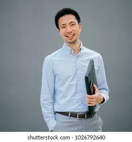 Portrait Of A Young, Chinese, Pan Asian Business Man Standing Against A Plain Grey Background And Smiling While He Holds His Leather Folio. He Is Well-dressed In Business Casual And Looks Relaxed. 