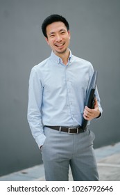 Portrait Of A Young, Chinese, Pan Asian Business Man Standing Against A Plain Grey Background And Smiling While He Holds His Leather Folio. He Is Well-dressed In Business Casual And Looks Relaxed. 