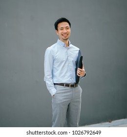 Portrait Of A Young, Chinese, Pan Asian Business Man Standing Against A Plain Grey Background And Smiling While He Holds His Leather Folio. He Is Well-dressed In Business Casual And Looks Relaxed. 