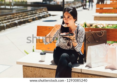 Portrait of young chinese woman eating dessert outdoors under sunshine sitting on bench chair with shopping bags beside in mall center stanford near the water pool. female lifestyle modern life.