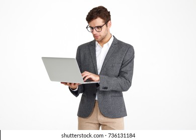 Portrait of a young concentrated businessman in eyeglasses and a jacket working on laptop computer while standing isolated over white background
