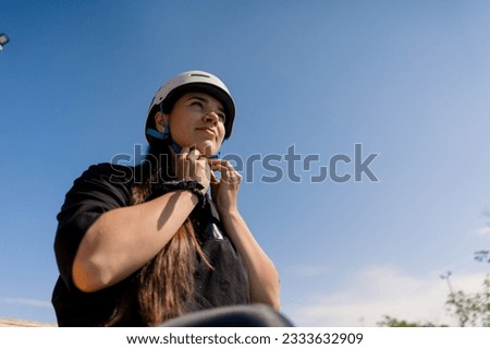 portrait of young concentrated hipster girl against sky background in skate park putting safety helmet before starting roller skating