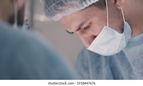 Portrait Of A Young Concentrated Male Doctor Or Assistant Wearing Medical Face Mask And Protective Cap. Action. Surgeon Looking Down With Calm Emotions During The Operation, Medicine Concept.