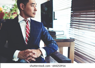 Portrait of young confident asian businessman with serious face sitting in modern office interior near big window, intelligent men entrepreneur in elegant suit thinking about something before meeting 