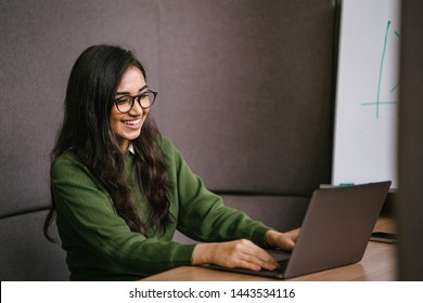 Portrait Of A Young, Confident And Attractive Indian Asian Student Working Or Studying On Her Laptop As She Sits In A Booth. She Is Dressed In A Preppy Sweater And Shirt And Glasses. 