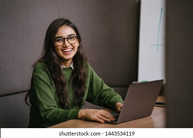 Portrait of a young, confident and attractive Indian Asian student working or studying on her laptop as she sits in a booth. She is dressed in a preppy sweater and shirt and glasses.  - Shutterstock ID 1443534119