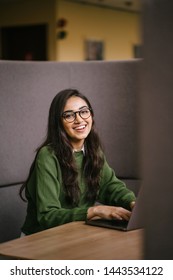 Portrait Of A Young, Confident And Attractive Indian Asian Student Working Or Studying On Her Laptop As She Sits In A Booth. She Is Dressed In A Preppy Sweater And Shirt And Glasses. 