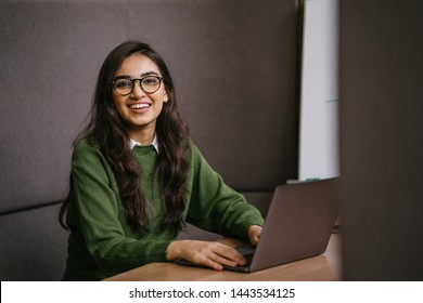 Portrait Of A Young, Confident And Attractive Indian Asian Student Working Or Studying On Her Laptop As She Sits In A Booth. She Is Dressed In A Preppy Sweater And Shirt And Glasses. 