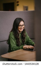 Portrait Of A Young, Confident And Attractive Indian Asian Student Working Or Studying On Her Laptop As She Sits In A Booth. She Is Dressed In A Preppy Sweater And Shirt And Glasses. 