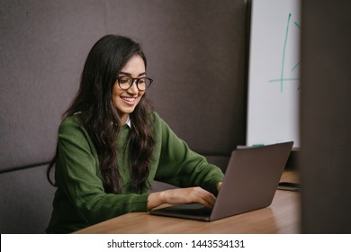 Portrait Of A Young, Confident And Attractive Indian Asian Student Working Or Studying On Her Laptop As She Sits In A Booth. She Is Dressed In A Preppy Sweater And Shirt And Glasses. 