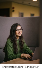 Portrait Of A Young, Confident And Attractive Indian Asian Student Working Or Studying On Her Laptop As She Sits In A Booth. She Is Dressed In A Preppy Sweater And Shirt And Glasses. 