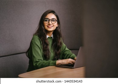 Portrait Of A Young, Confident And Attractive Indian Asian Student Working Or Studying On Her Laptop As She Sits In A Booth. She Is Dressed In A Preppy Sweater And Shirt And Glasses. 