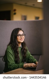 Portrait Of A Young, Confident And Attractive Indian Asian Student Working Or Studying On Her Laptop As She Sits In A Booth. She Is Dressed In A Preppy Sweater And Shirt And Glasses. 