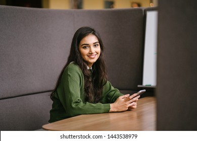 Portrait Of A Young, Confident, Intelligent-looking And Attractive Indian Asian Woman Using Her Smartphone. She Is Wearing A Preppy Green Sweater And Smiling Happily. 