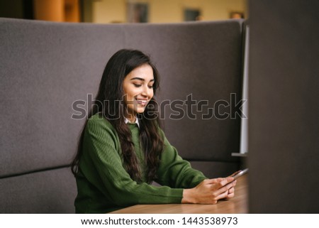 Portrait of a young, confident, intelligent-looking and attractive Indian Asian woman using her smartphone. She is wearing a preppy green sweater and smiling happily. 