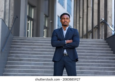 Portrait Young Confident Successful African American Businessman Looking At Camera Serious Business Man Standing On Urban Background Of Modern Office Building Outdoors. Male A Formal Suit Arms Crossed