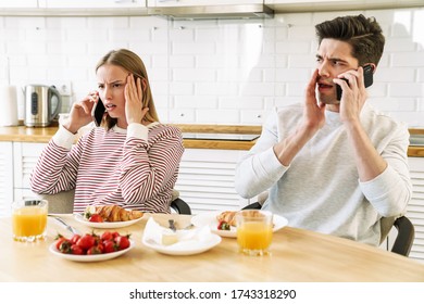 Portrait Of Young Confused Couple Talking On Smartphones While Having Breakfast In Cozy Kitchen At Home