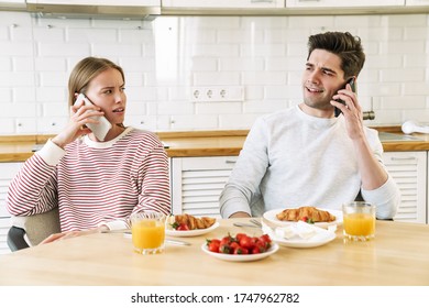 Portrait Of Young Confused Couple Talking On Smartphones While Having Breakfast In Cozy Kitchen At Home