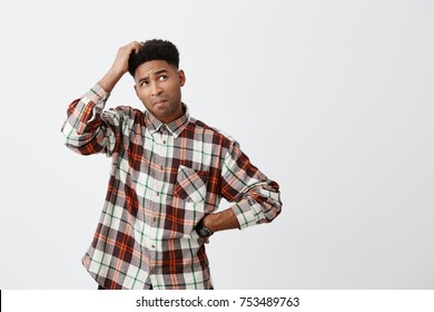 Portrait of young confused dark-skinned handsome man with afro hairstyle in checkered shirt holding head with hand, looking aside with satisfied expression, don't know what to do with debts