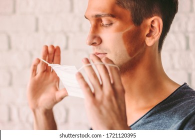 Portrait Young Confused Hipster Man With Funny Sun Tanned Lines On Face Holding Protective Mask In Hands. Angry Man Patient With Sunburn Tan Skin After Wearing Medical Face Mask. COVID-19 Prevention