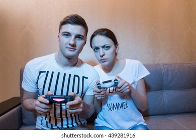 Portrait Of Young Couple Of Boy And Girl, With Very Serious And Pensive Face, Playing Video Game Sitting On The Sofa In Front Of TV At Home In A Relaxed Atmosphere