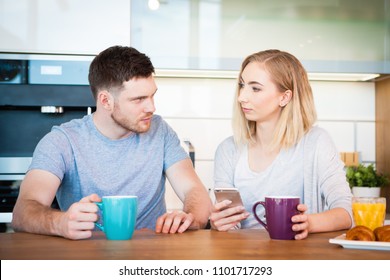 Portrait of a young couple having breakfast while sitting at the table in a kitchen at home and having quarrel because of smartphone