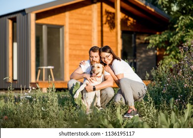 Portrait Of A Young Couple Hugging Happy Dog Sitting On The Backyard Of Their Wooden Country House