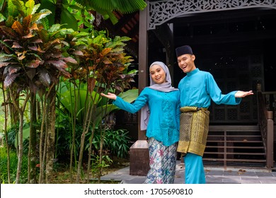 A portrait of young couple of malay muslim in traditional costume during Aidilfitri celebration showing welcome greeting gesture by traditional wooden house. Raya and Muslim fashion attire concept.