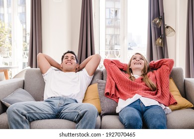 Portrait Of Young Couple Putting Hands Behind Head And Relaxing While Sitting On Sofa At Home.