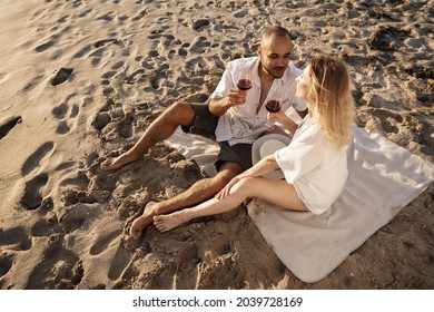 Portrait Of A Young Couple Sitting On The Beach And Drinking Wine