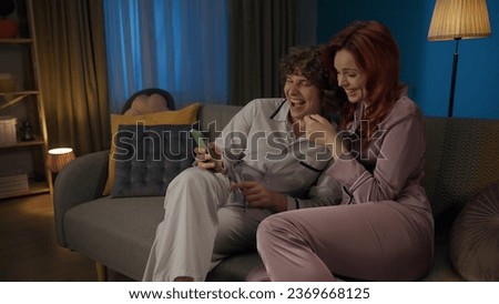 Portrait of young couple spending time together. Man and woman in pajamas sitting on the sofa, holding smartphone laughing.