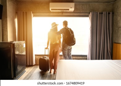 Portrait Of Young Couple Tourist Standing Nearly Window, Looking To Beautiful View Outside In Hotel/resort Bedroom After Check-in. Conceptual Of Couple Travel And Vacation In Their Honeymoon Period.