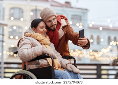 Portrait of young couple with woman using wheelchair taking selfie photo outdoors in winter and using smartphone - Powered by Shutterstock