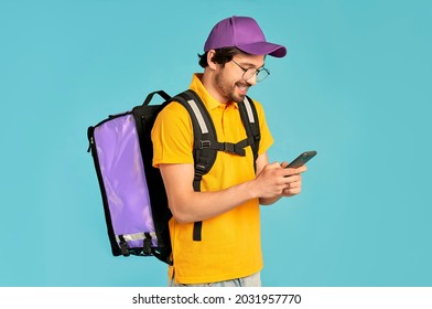 Portrait Of A Young Courier, Delivery Man In Uniform With A Thermo Backpack And A Smartphone Isolated On A Blue Background. Fast Home Delivery. Online Order.