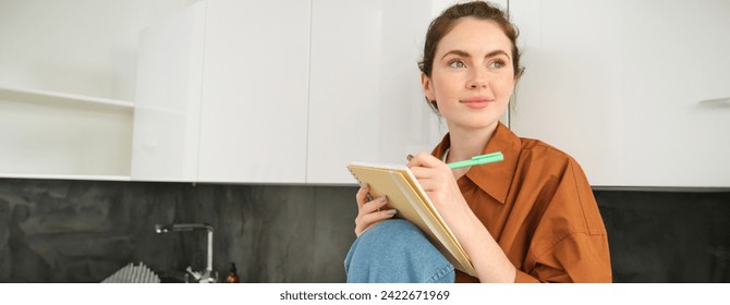 Portrait of young creative girl, writing down her ideas in planner, does homework in notebook, sits at home on kitchen counter. - Powered by Shutterstock