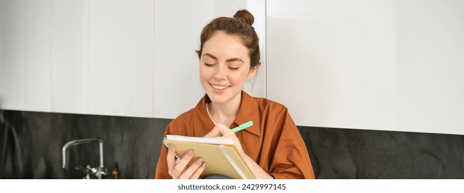 Portrait of young creative girl, writing down her ideas in planner, does homework in notebook, sits at home on kitchen counter. - Powered by Shutterstock