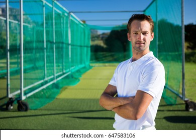 Portrait of young cricketer with arms crossed standing against net on field