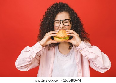 Portrait Of Young Curly Beautiful Hungry Woman Eating Burger. Isolated Portrait Of Student With Fast Food Over Red Background. Diet Concept. 