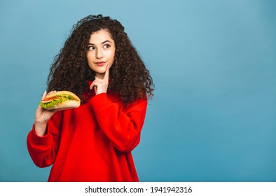 Portrait Of Young Curly Beautiful Hungry Woman Eating Burger. Isolated Portrait Of Student With Fast Food Over Blue Background. Diet Concept. 