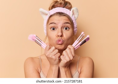 Portrait of young cute European female model going to apply makeup after taking shower holds cosmetic brushes near face keeps lips folded wears headband has combed hair poses indoor at studio