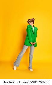 Portrait of young cute girl in bright green jacket posing over yellow studio background. Feeling shy. Concept of youth, beauty, casual fashion, lifestyle, emotions, facial expression. Ad - Shutterstock ID 2281703905