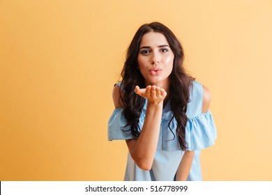 Portrait of a young cute woman in blue dress blowing kiss at camera isolated over orange background