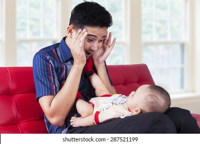 Portrait Of Young Dad Sitting On Sofa At Home While Playing Peekaboo With His Baby Boy