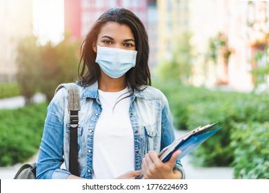 Portrait Of Young Dark Skinned Female Student Wearing Medical Mask Posing Outdoors With A Backpack And Pile Of Books.