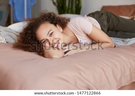 Portrait of young dark skinned happy lady lying on the bed in she's room and enjoy the sunny morning at home, broadly smiling and looking at the camera.