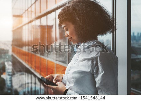 Portrait of a young dazzling African-American businesswoman standing near a panoramic window of a modern luxurious office high-rise and using her smartphone; a copy space area on the left for your ad