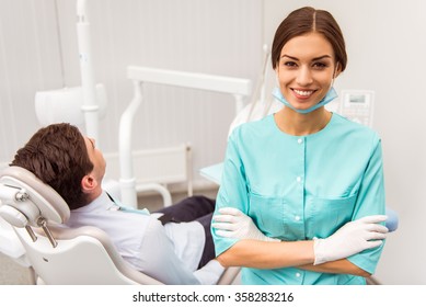 Portrait Of A Young Dentist With Folded Hands, Against The Background Of A Patient  In The Dental Chair