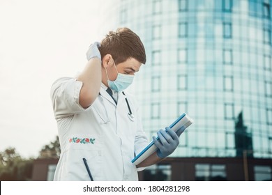 Portrait Of Young Doctor Standing Outside And Scratching The Back Of His Head. Man Wear Medical Gown, Blue Face Mask, Stethoscope And Gloves. Coronavirus Concept. Image With Copy Space.