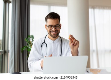 Portrait Of Young Doctor Talking To Online Patient On Laptop Screen Sitting At Clinic Office Desk Giving Online Consultation For Domestic Health Treatment. Telemedicine Remote Medical Appointment